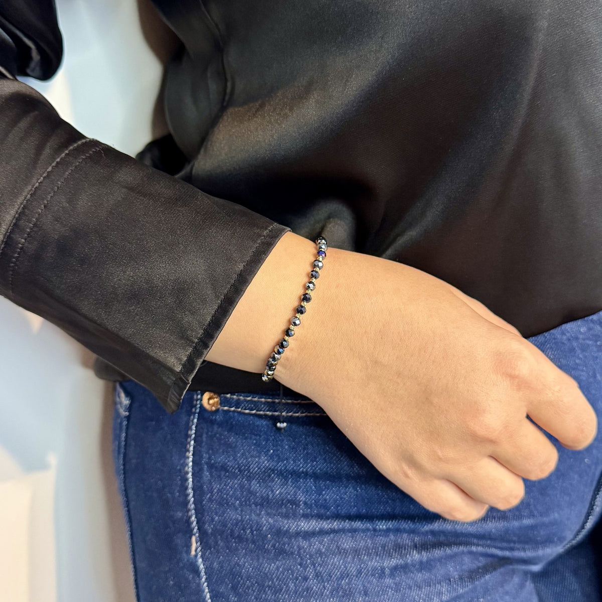 Brățara ZERAH din hematit iridescent și aur 14k purtată pe încheietura unei femei, lângă mâneca unei bluze negre din satin #Culoare_hematit iridescent
